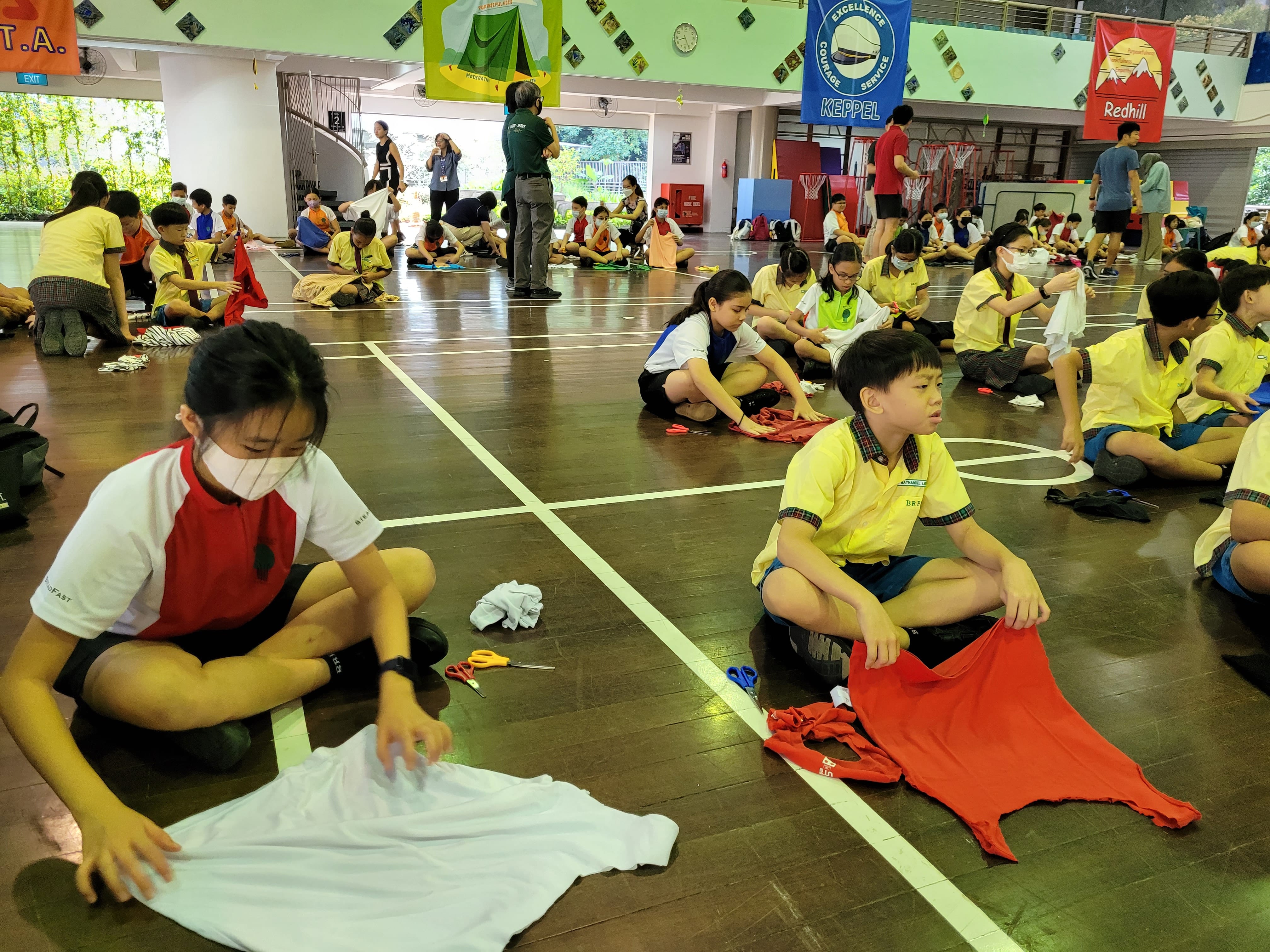 Students sit on a gym floor cutting and folding T-shirts, with scissors and fabric scattered around.
